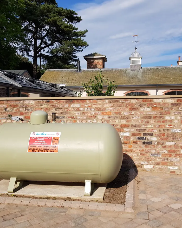 An above-ground LPG tank on a paved base beside the brick wall of a country property