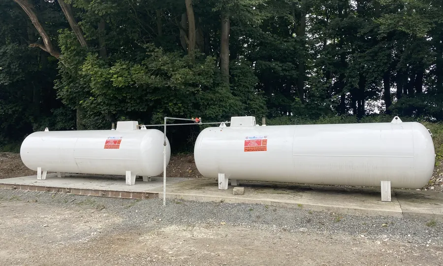 Two white above-ground bulk LPG tanks on a gravel base at the edge of woodland