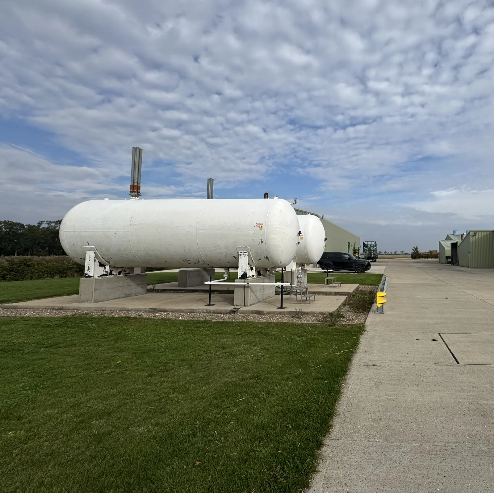 Two large white above-ground LPG tanks on a concrete base beside a green farm building under a broken-cloud sky