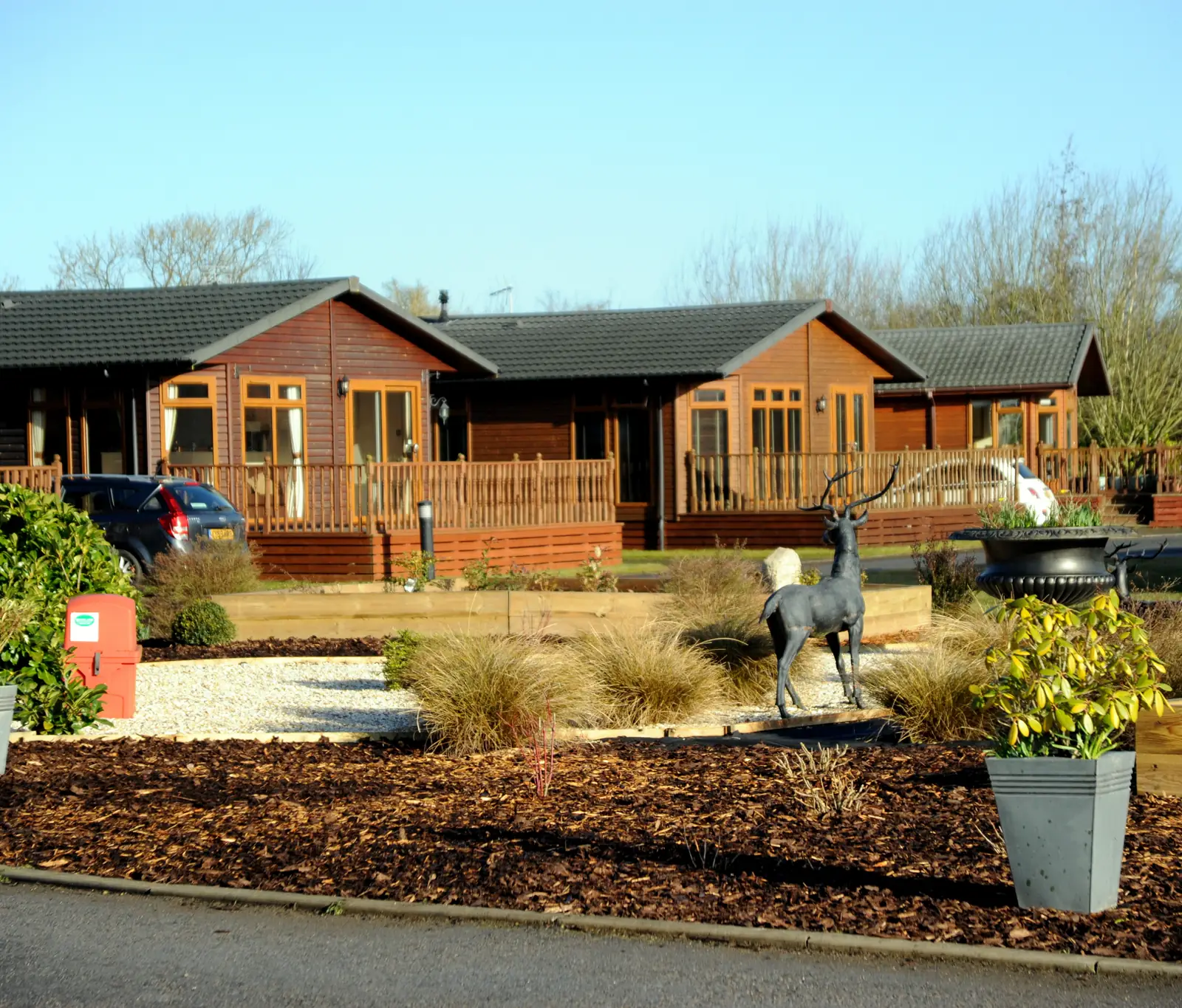 Timber holiday lodges with decking behind a landscaped garden and a bronze stag sculpture at a holiday park
